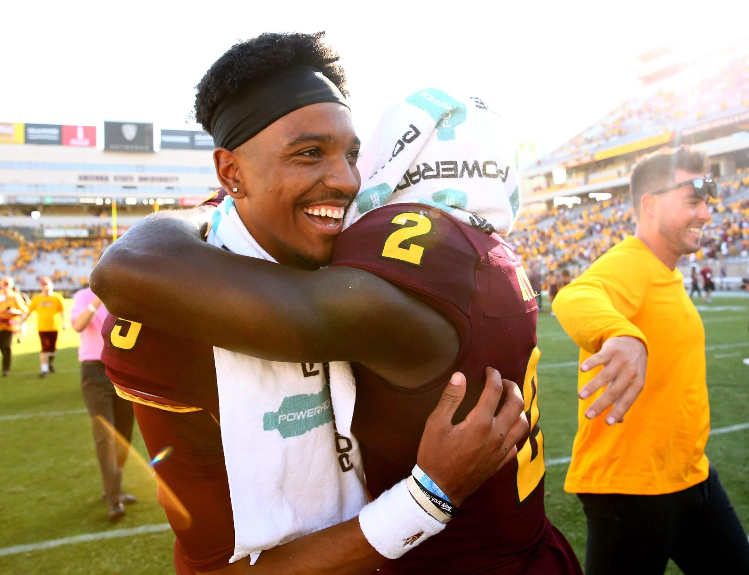Brandon Aiyuk, in a maroon No. 2 Arizona State uniform with a towel over his head, hugs Jayden Daniels, who is wearing a maroon No. 5 uniform and smiling wide.
