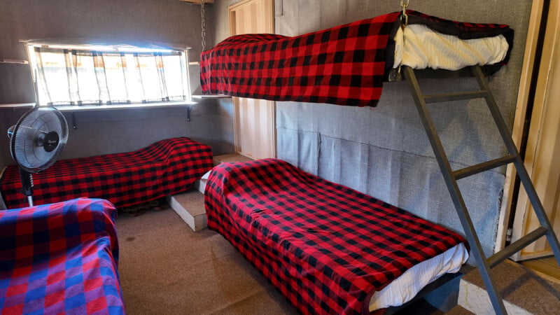 A small room with three single beds covered in red and black checkered blankets, a metal bunk bed ladder, a standing fan, and a window with bars letting in sunlight.