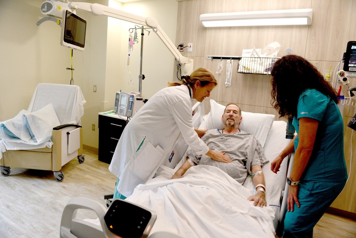 A doctor and nurse examine a male patient lying in a hospital bed in a modern, well-lit hospital room.