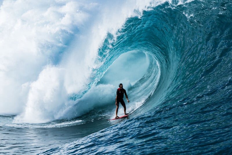 A surfer in a black wetsuit rides a large, powerful blue wave, positioned inside the barrel as water crashes dramatically around him.