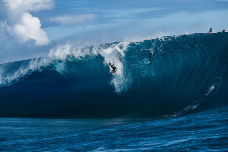 A surfer rides down the face of a massive, barreling blue wave under a clear sky, with the ocean and a few onlookers in the background.