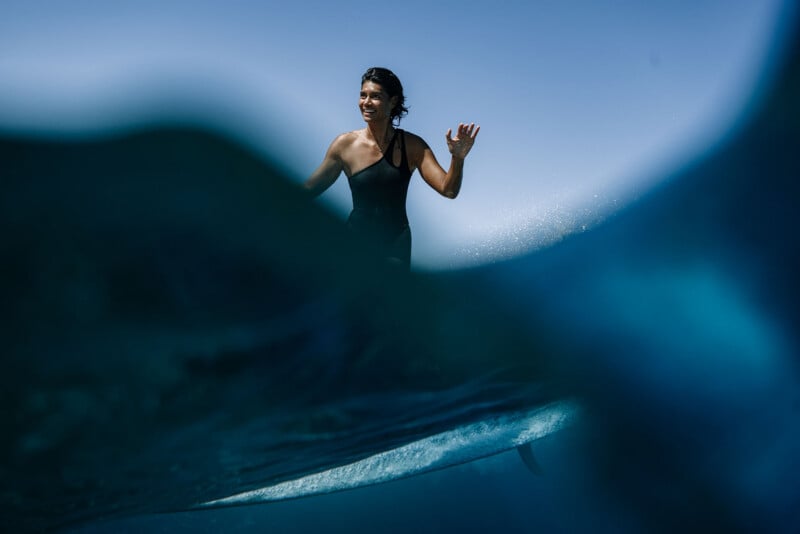 A person in a black swimsuit stands on a surfboard in the ocean, partially obscured by a wave, with a clear blue sky in the background.