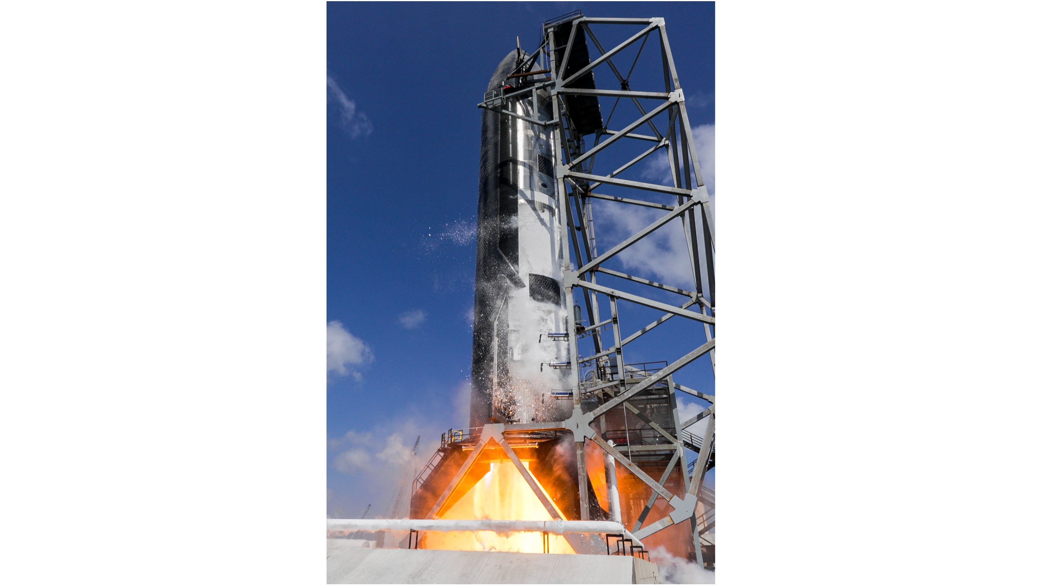 a dark, tall, conical spacecraft fires its engines on a test stand beneath a blue but cloudy sky