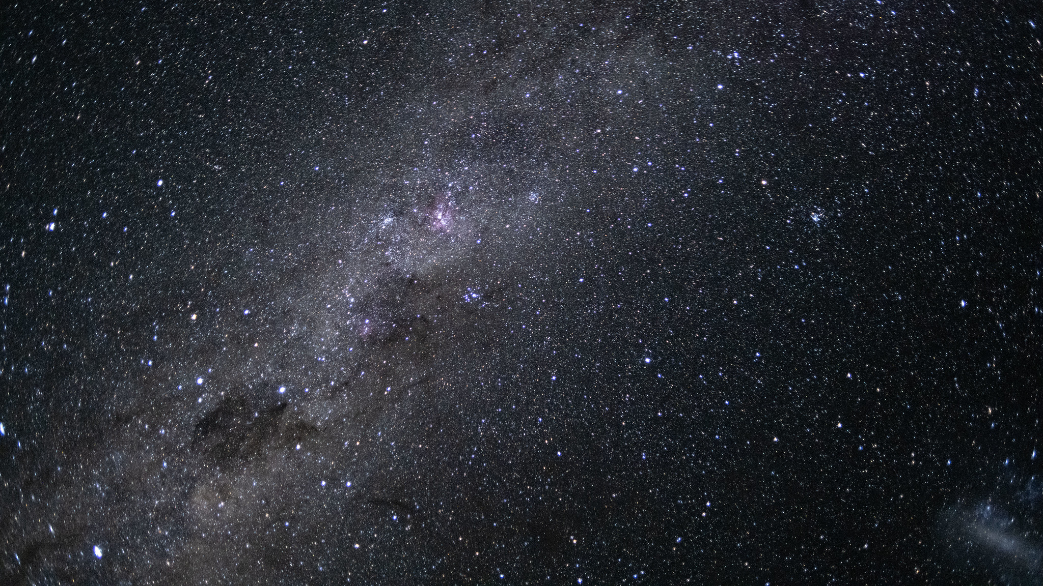 The glowing band of the Milky Way can be seen streaking across an image captured from near lunar space, from the bottom left to top right of the photo. Pink nebulas and dark dusty clouds can be seen threading the galactic plane, and a satellite galaxy is visible as a smudge of white light to the bottom right of the screen.