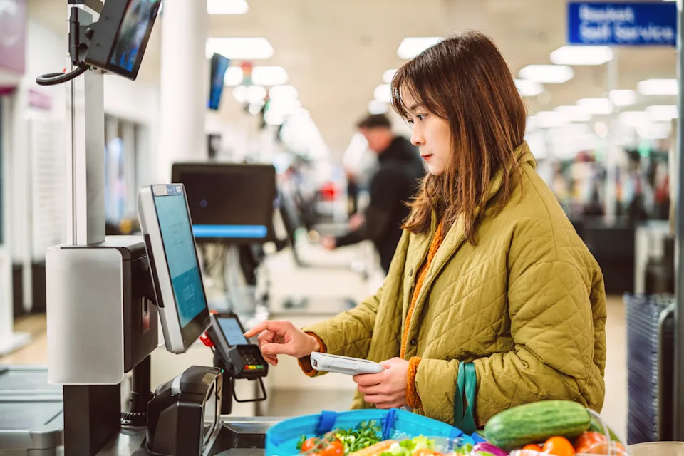Asian woman using a self checkout machine in a supermarket, making a contactless payment during grocery shopping. Concept of mobile payment, digital wallets, retail technology, and cashless society.