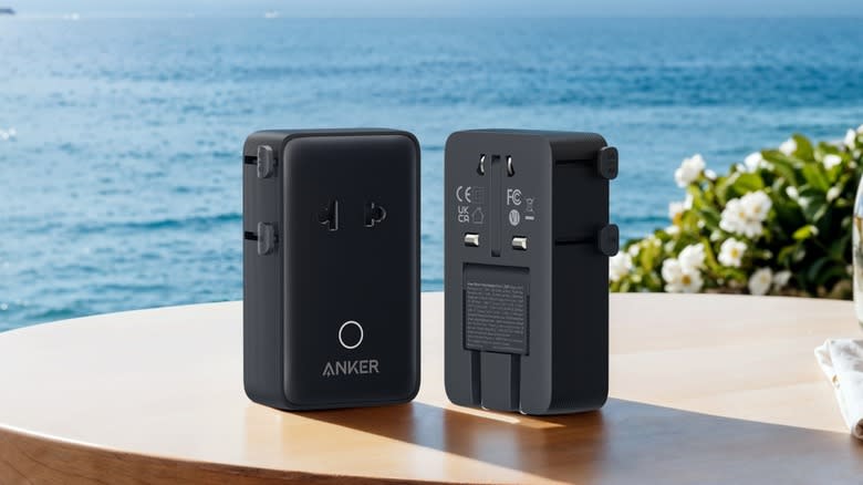 A pair of Anker Nano travel adapters viewed from the front and back, on top of a wooden table with the ocean in the background.