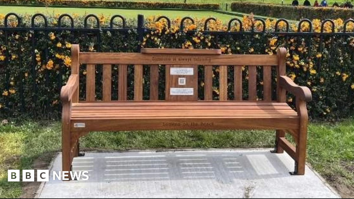 A bench placed on a concrete slab. It is placed in front of a metal fence.
