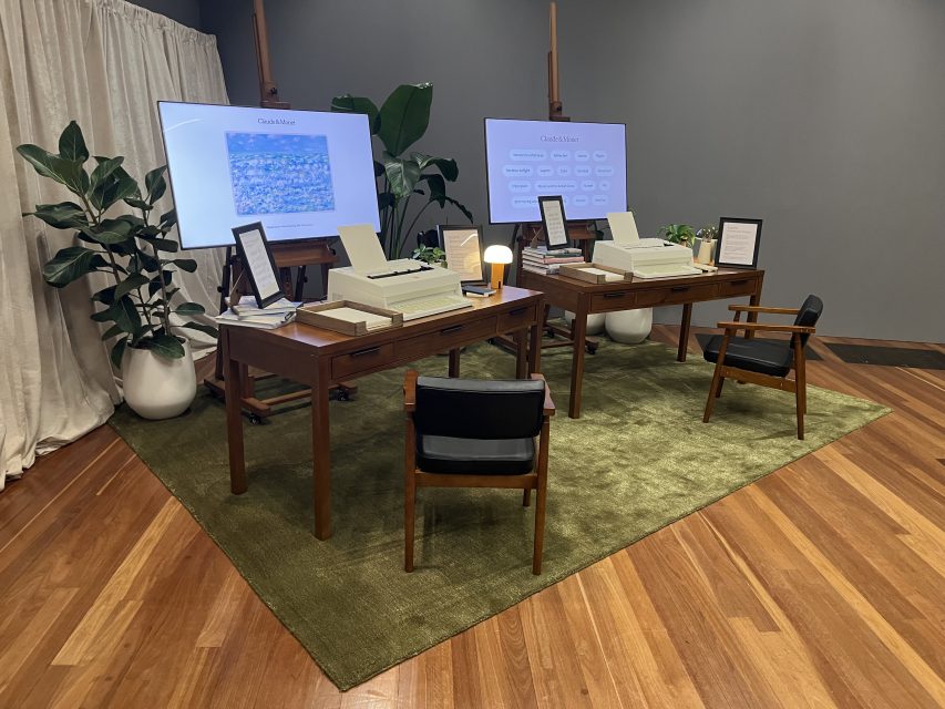 Two vintage computer workstations with old monitors and keyboards are set up on a green rug, surrounded by plants and two chairs, in a modern exhibit space.