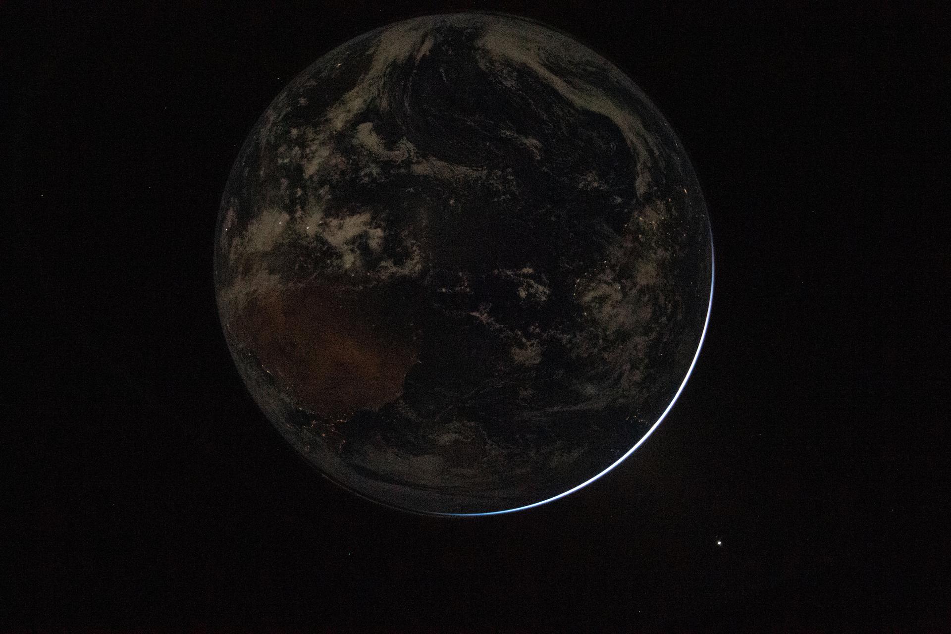 A view of the Earth from space, mostly in shadow with a thin crescent of sunlight illuminating the edge, evokes the hope and wonder inspired by missions like Artemis II against a vast, dark background.