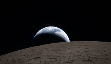 A muted blue Earth with bright white clouds sets behind the cratered lunar surface. The dark portion of Earth is experiencing nighttime. On Earth’s day side, swirling clouds are visible over the Australia and Oceania region. In the foreground, Ohm crater has terraced edges and a flat floor interrupted by central peaks. Central peaks form in complex craters when the lunar surface, liquefied on impact, splashes upwards during the crater’s formation.
