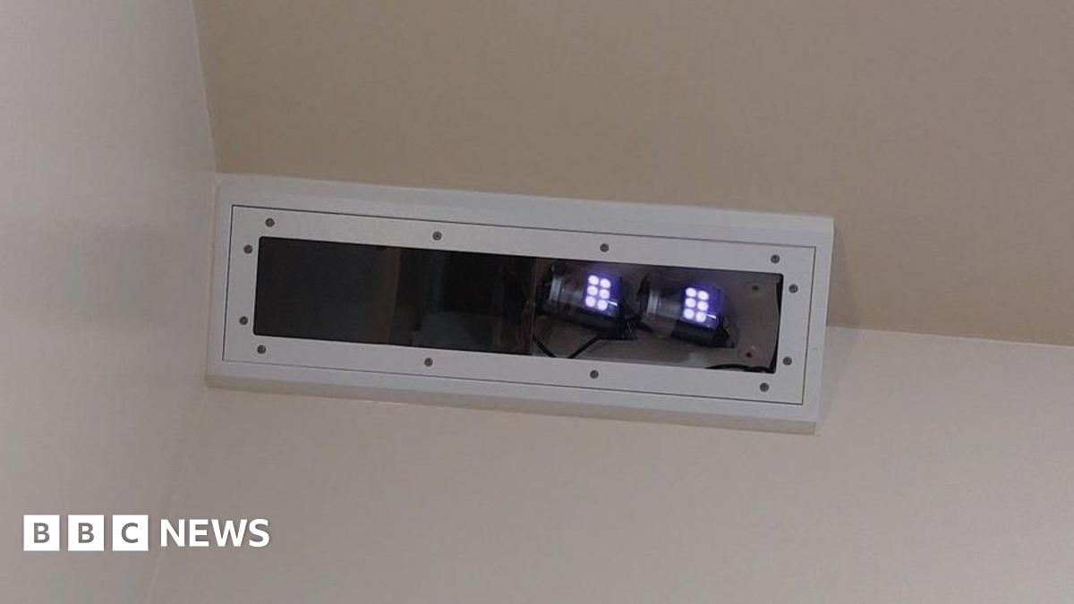 The Oxevision technology - which is an infrared camera - placed near the ceiling in the corner of a bedroom. It is in a white rectangle box and small white lights can be seen at one end.