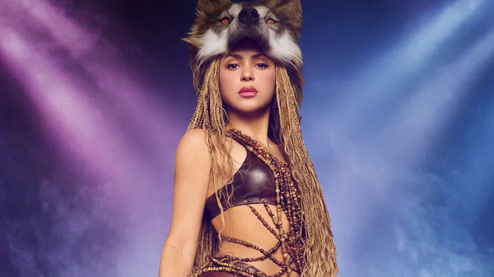 Shakira wearing a wolf headdress and braided outfit stands confidently against a colorful, smoky background.