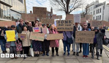 A group of people stand outside a building on a street. They are holding home-made placards in support of a mental health service which risks being cut.