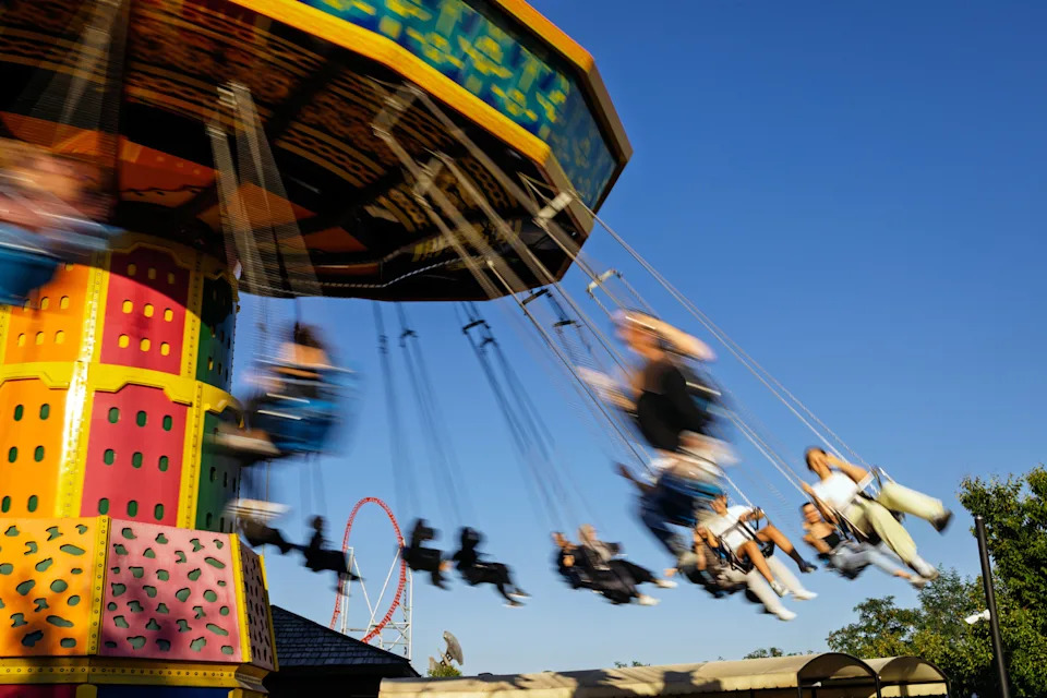 People enjoy a spinning swing ride at an amusement park, with blurred motion capturing the thrill. A roller coaster is visible in the background
