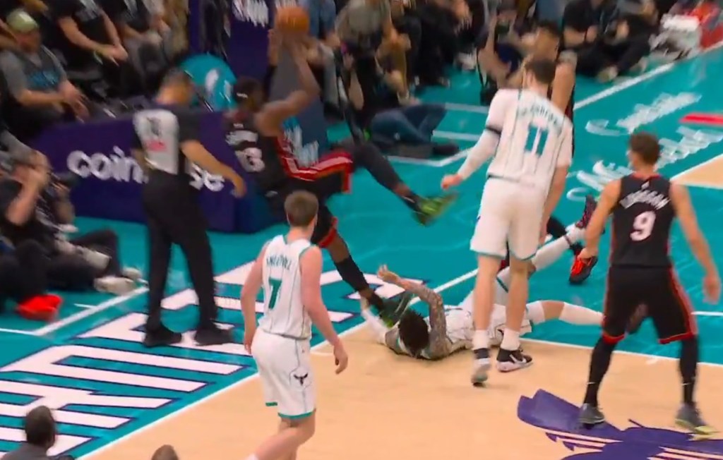 A basketball player in a black and red uniform holding a basketball, jumping over another player in a white and green uniform who has fallen to the ground.