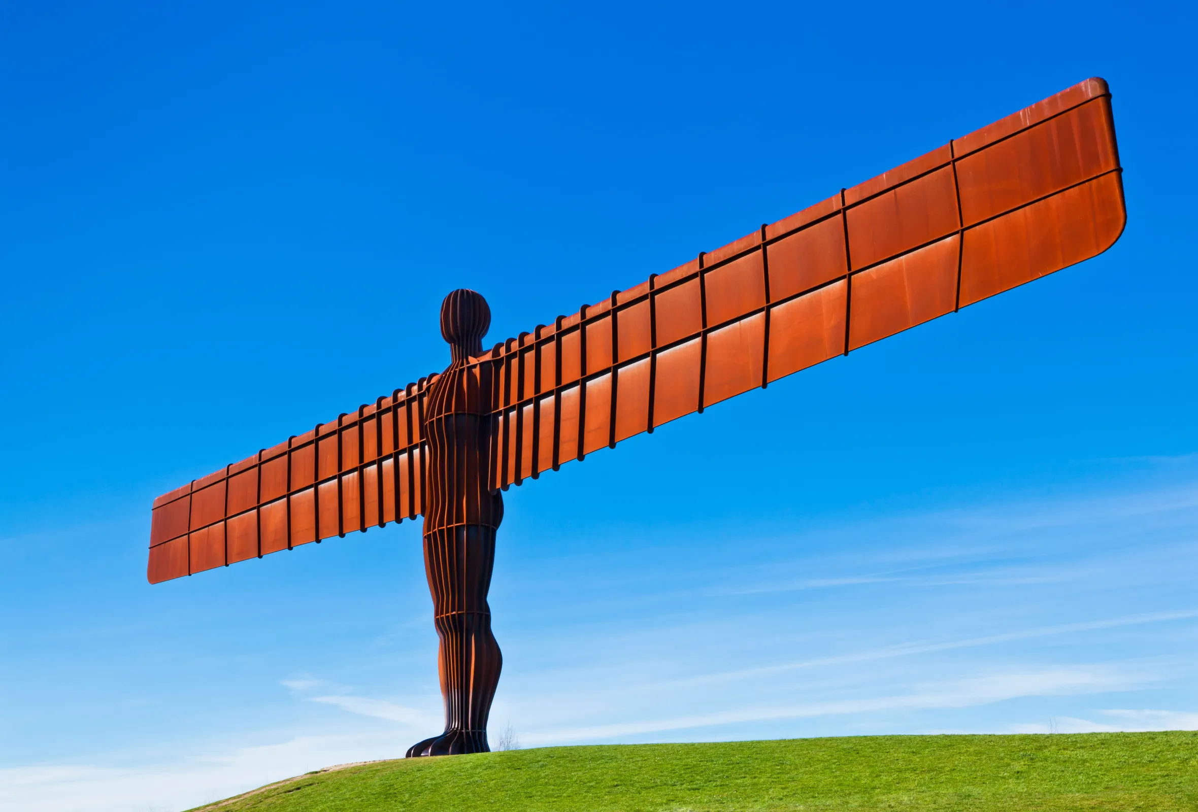 The Angel of the North, a rust-colored steel sculpture by Antony Gormley, stands on a grassy hill against a clear blue sky.