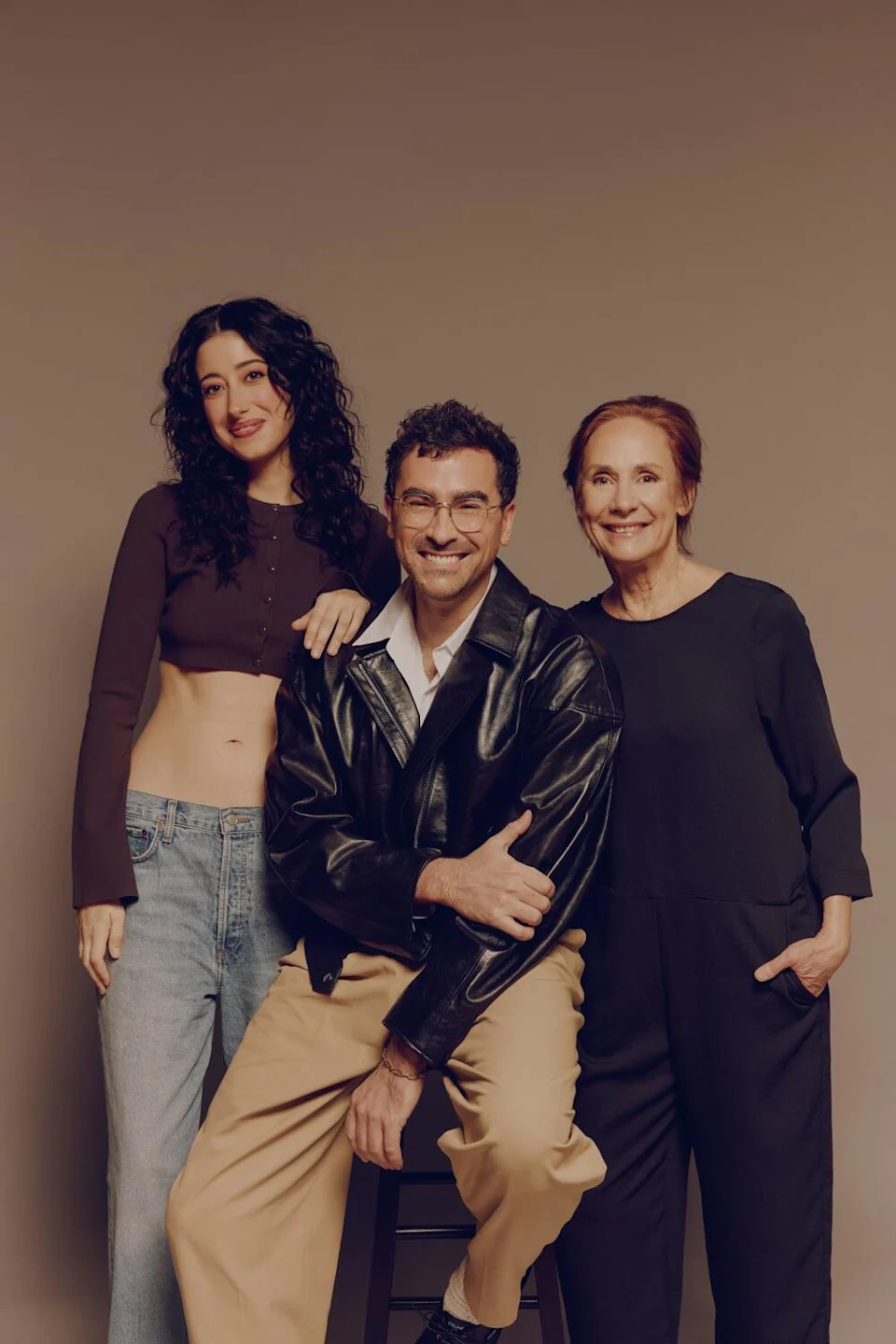 Two women flank a smiling man in a black leather jacket seated on a stool.