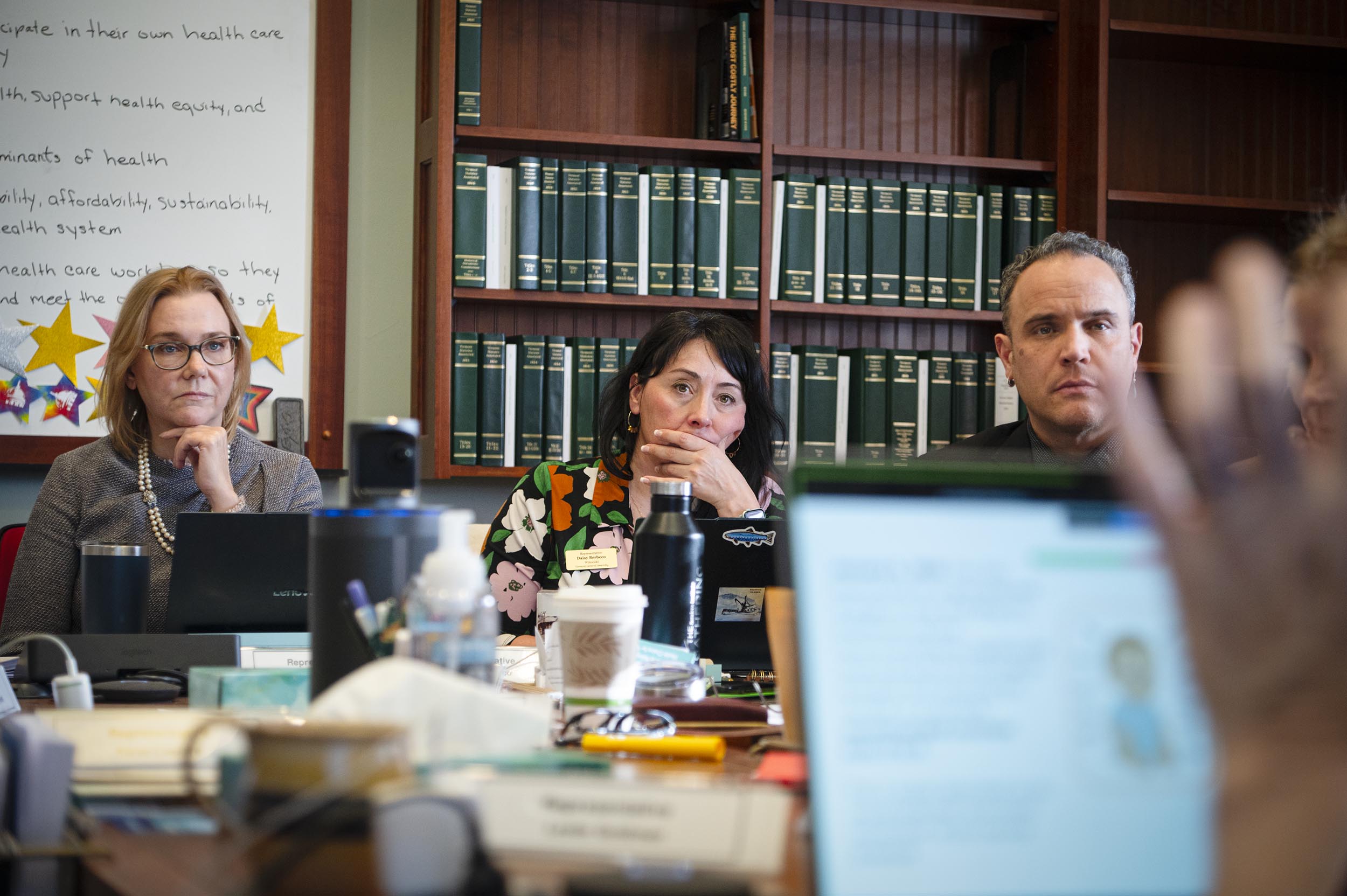 Three people sit at a conference table, listening attentively during a meeting, with bookshelves and a whiteboard in the background.