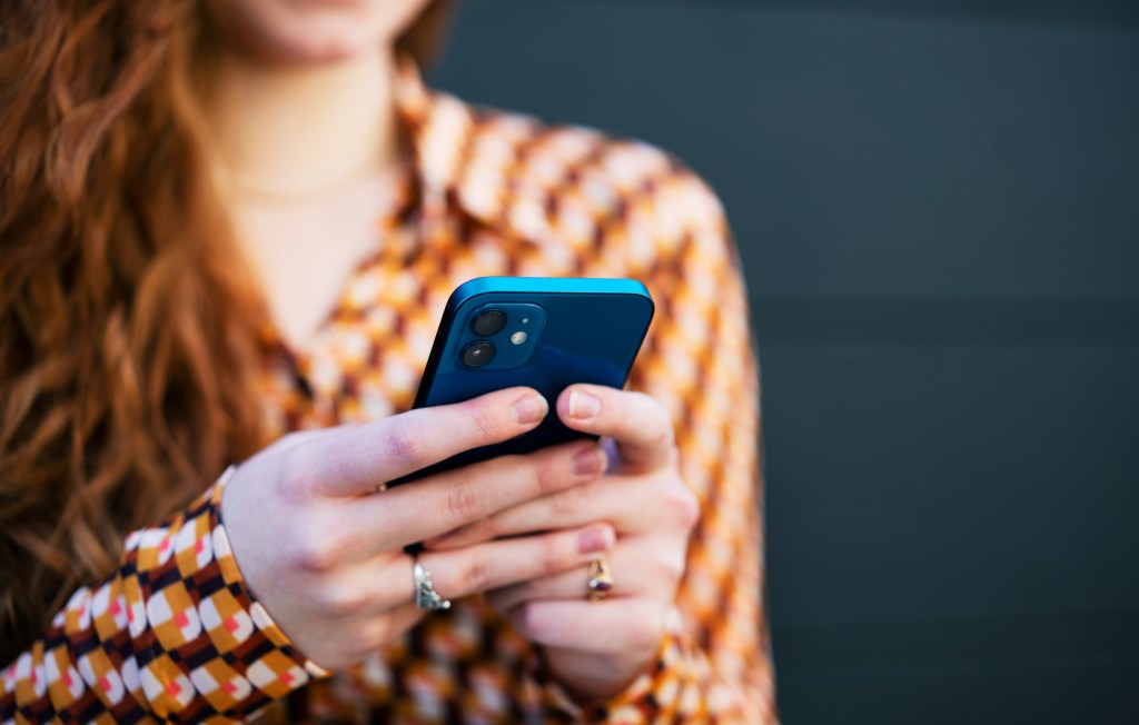 A close-up of a woman's hands holding a blue smartphone, with her orange patterned shirt and red hair blurred in the background.