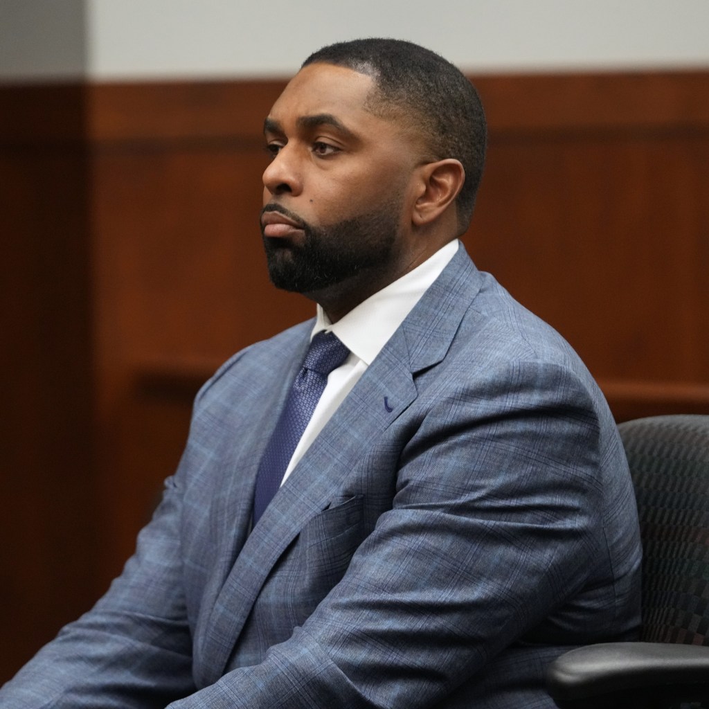 Sherrone Moore, former Michigan football coach, wearing a blue plaid suit and a blue patterned tie, sits in a chair in court.