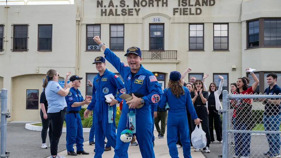 a man in an orange flight suit smiles while carrying a small stuffed moon