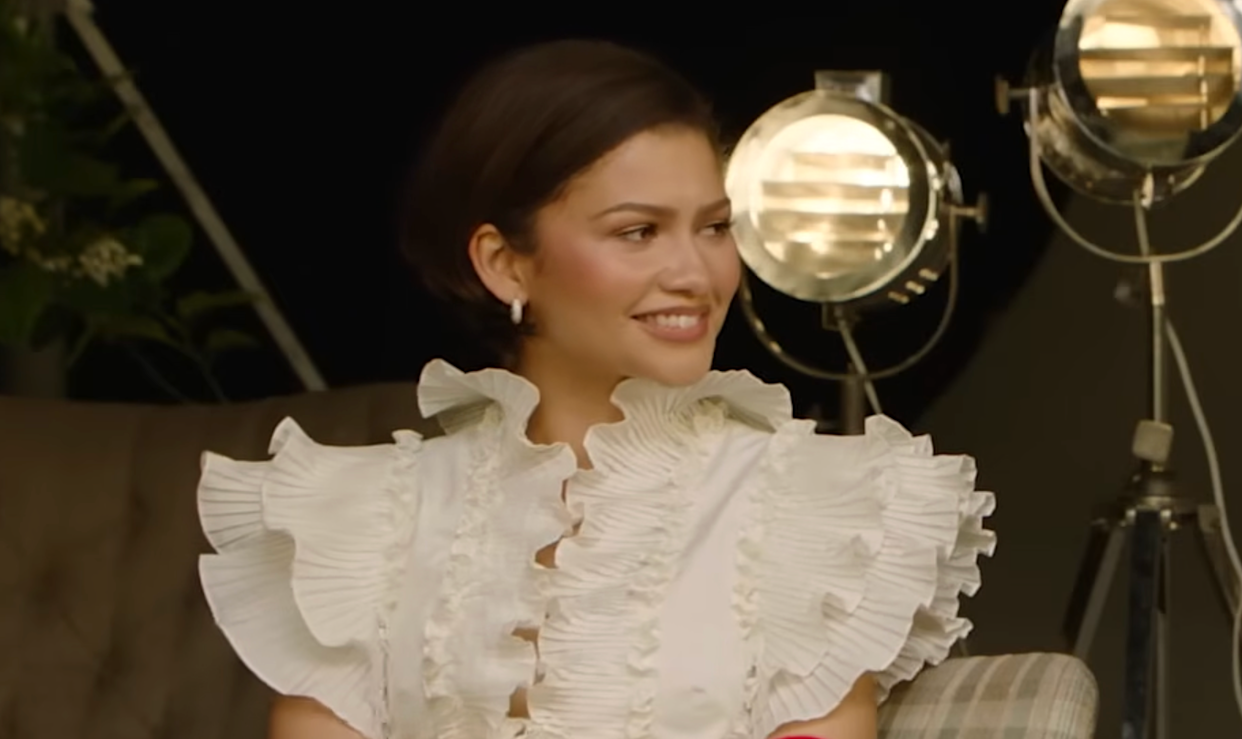 Person in white ruffled attire sitting on a couch with studio lights in the background.