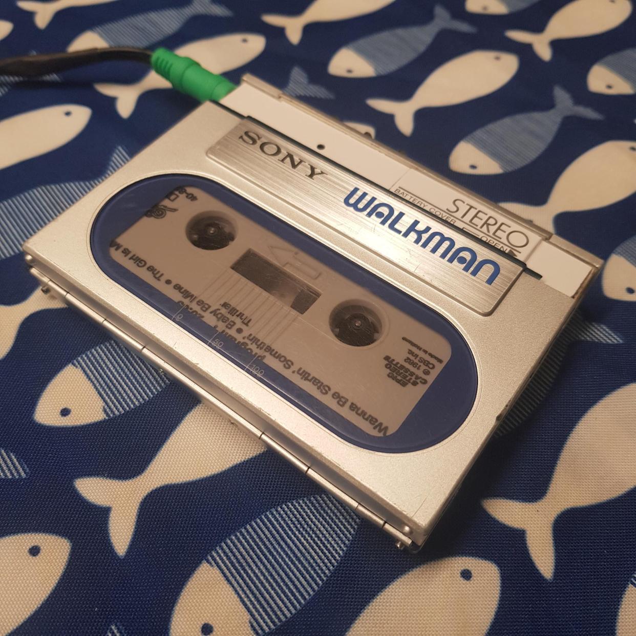 A silver Sony Walkman cassette player with a visible cassette tape inside rests on a blue and white fish-patterned fabric. A green audio jack is plugged into the Walkman.