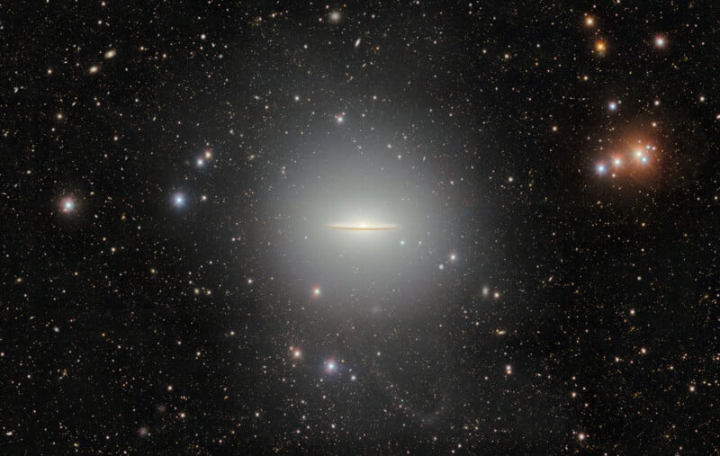 A bright, edge-on galaxy, known as the Sombrero Galaxy, appears in the center of a star-filled sky with a glowing halo surrounding it. Numerous stars and distant galaxies are scattered throughout the background.