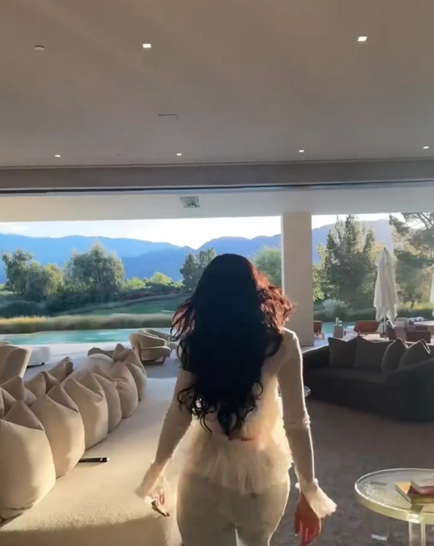 Kylie Jenner in a chic, fluffy outfit with long hair, walks through modern living room towards a scenic window view of mountains and a serene lake
