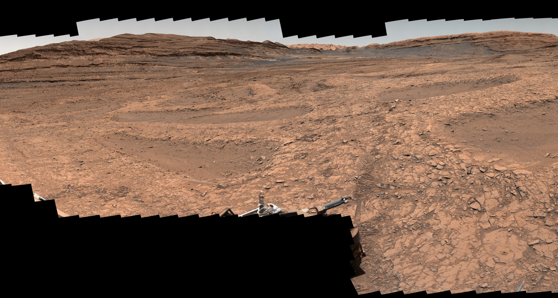 A series of shallow, sand-filled pits with low ridges spread across a tawny Martian landscape. Rover tracks stretch toward the horizon at left, and steep ridgetops loom in the background.