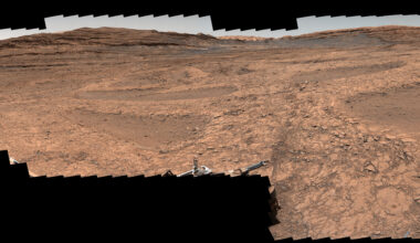 A series of shallow, sand-filled pits with low ridges spread across a tawny Martian landscape. Rover tracks stretch toward the horizon at left, and steep ridgetops loom in the background.