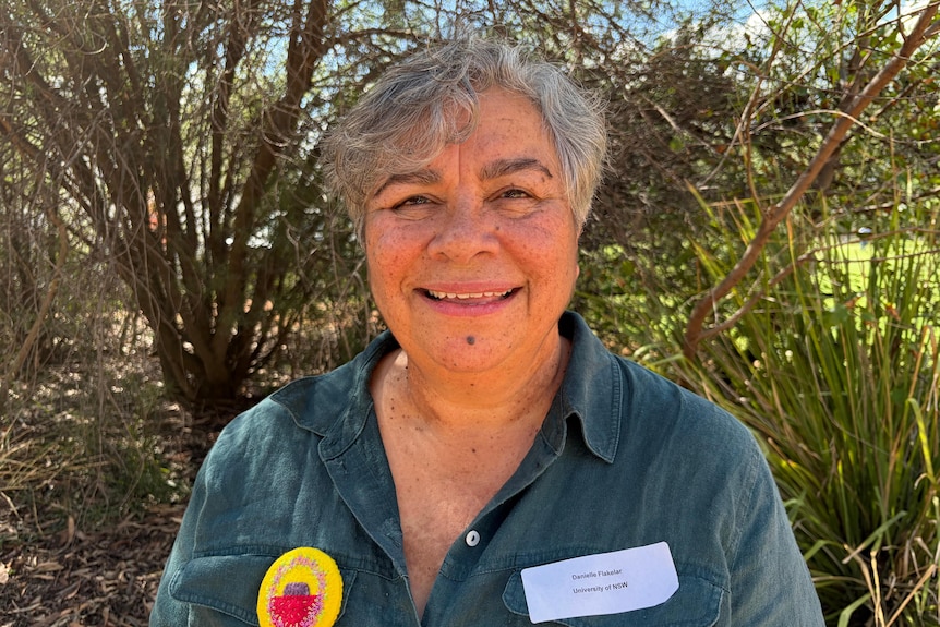 A First Nations woman smiles at the camera with greenery behind her