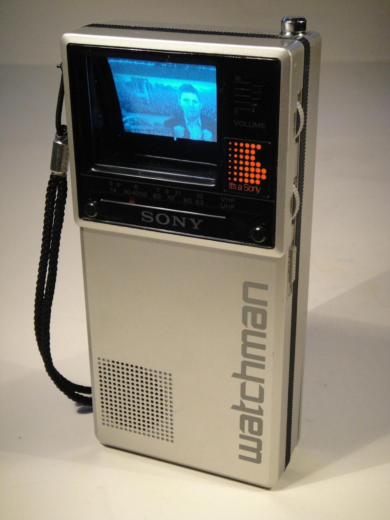 A vintage silver Sony Watchman portable TV displays a black-and-white image on its small screen, with buttons, a volume dial, a speaker, and a wrist strap visible on the side.