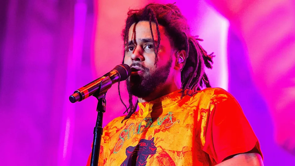 J. Cole performing on stage, wearing a vibrant orange shirt, with a microphone against a colorful background.