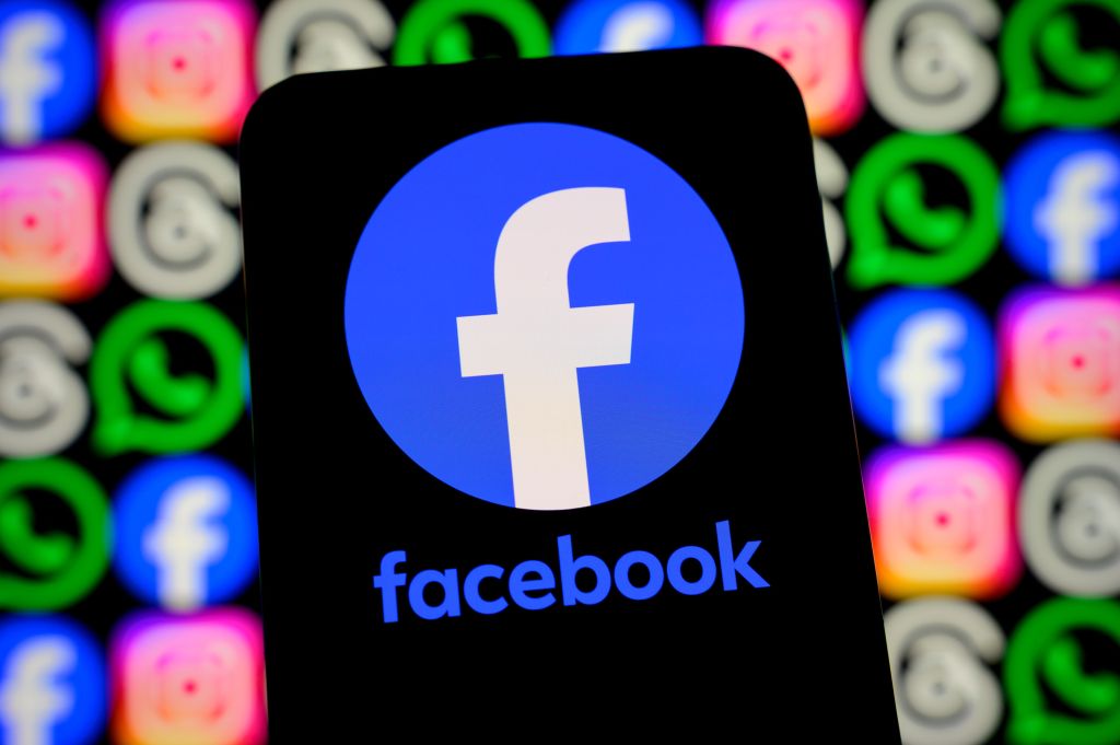 A close-up of a smartphone screen displaying the Facebook logo and name, with other blurred social media app icons in the background.
