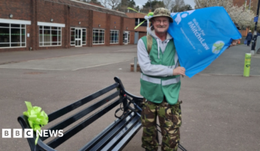 A man wearing a green high-vis jacket, a grey hoodie and camouflage trousers and a hat, holding a blue flag reading "Walking for Mitchs Law" stands next to a black bench with a green bow on it. There is a building behind him