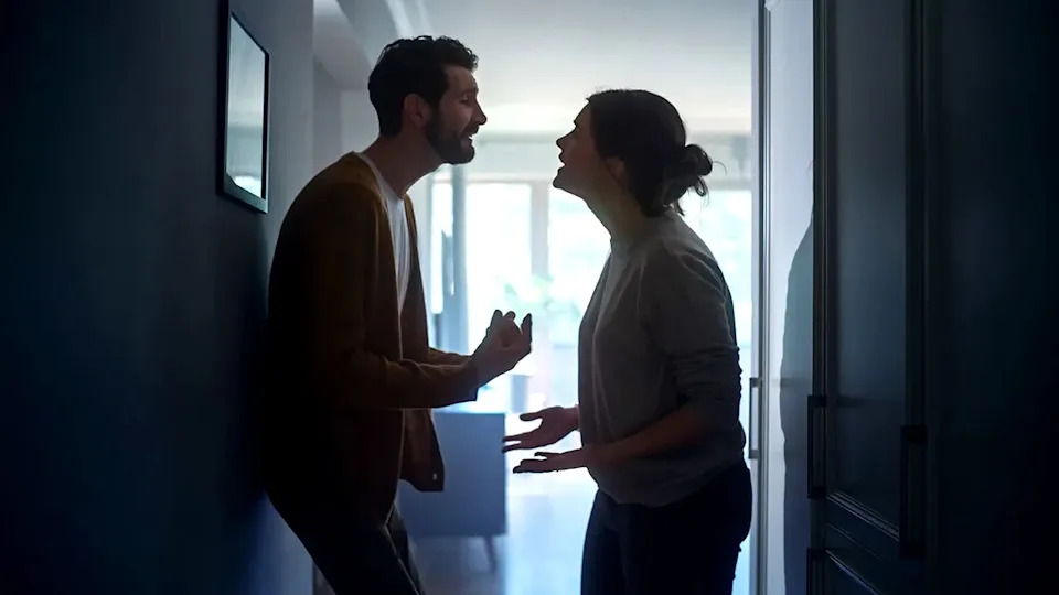 A young woman and man arguing in a dark apartment hallway