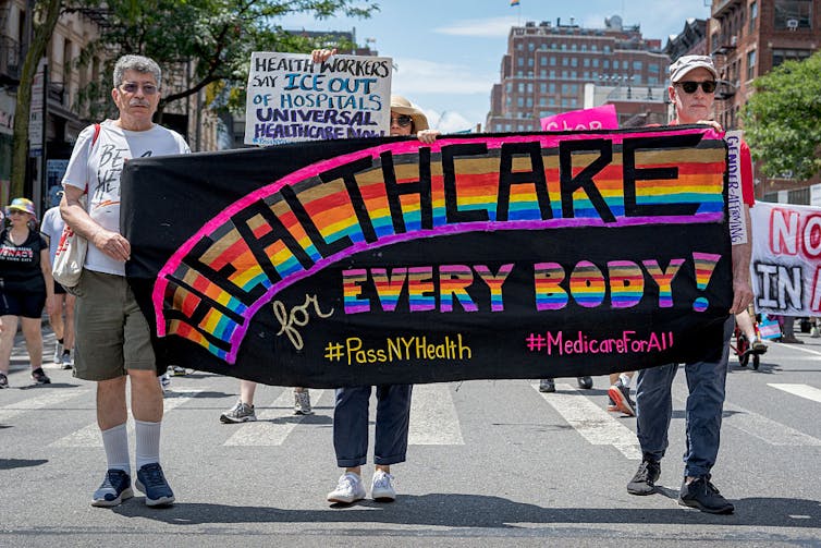 Marchers holding sign reading 'HEALTHCARE for EVERY BODY!' in rainbow coloring
