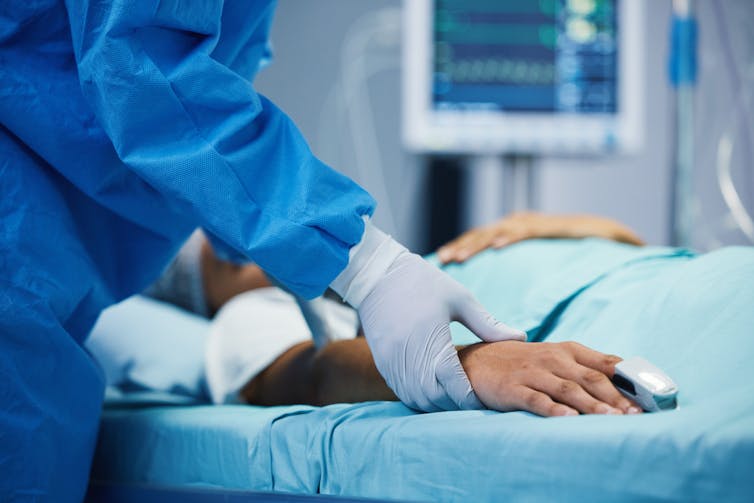 Clinician in scrubs and gloves holding wrist of patient lying on a hospital bed