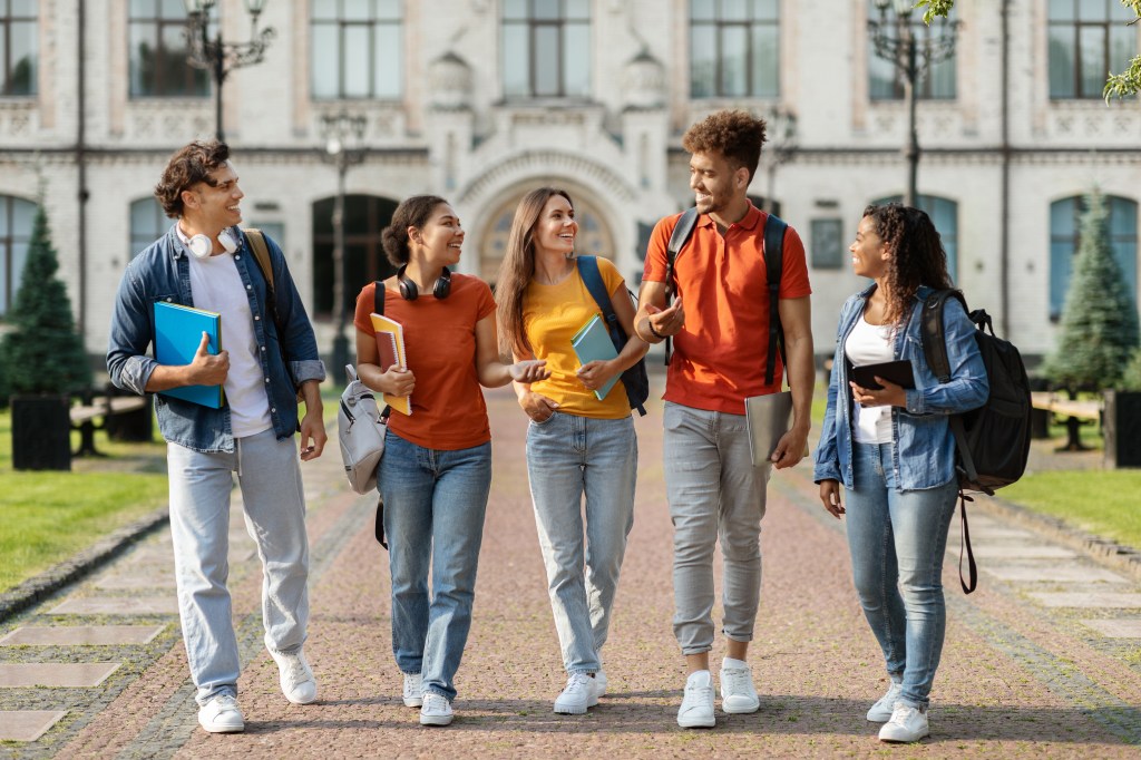 Five happy university students walking together on campus, chatting and laughing.