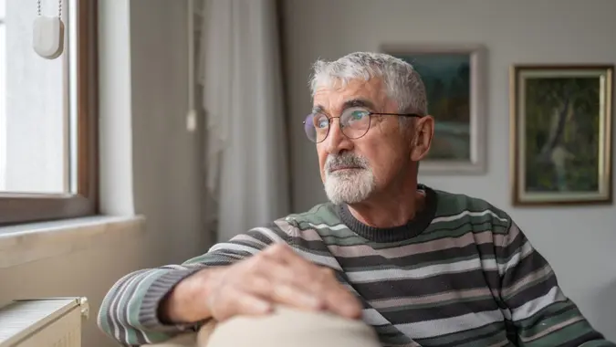 Senior Man Sitting On Sofa By Window And Looking Outside Through Window At Home.