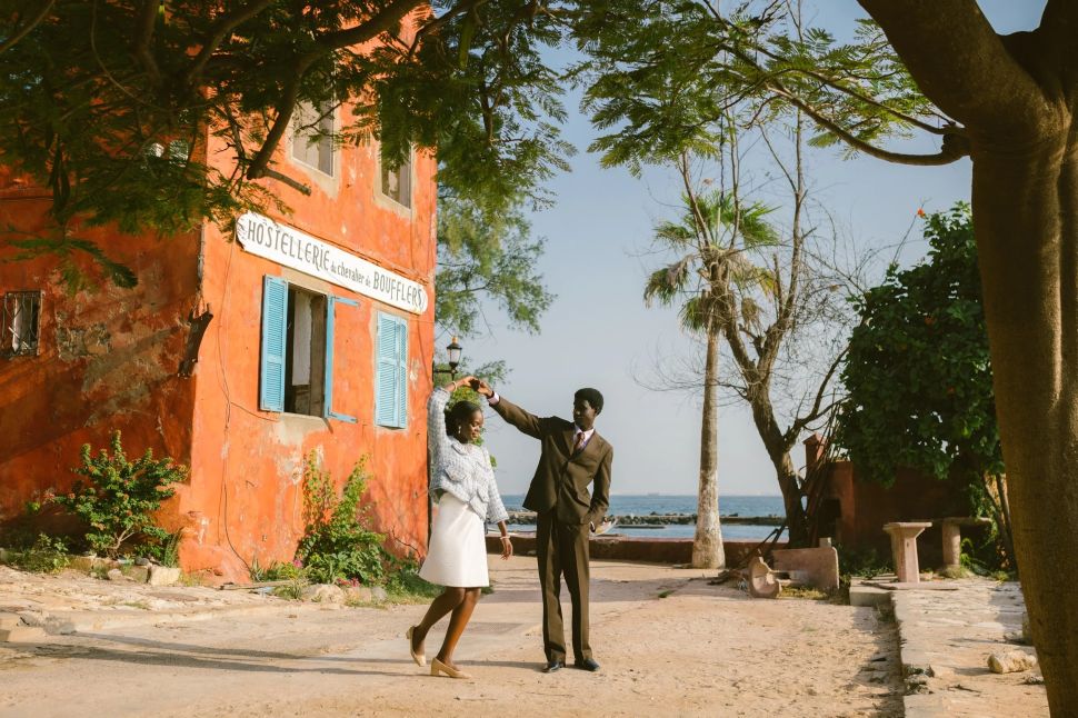 A couple dances outdoors near a vivid orange building labeled “Hostellerie du Chevalier de Boufflers,” with trees framing a coastal view in the background.
