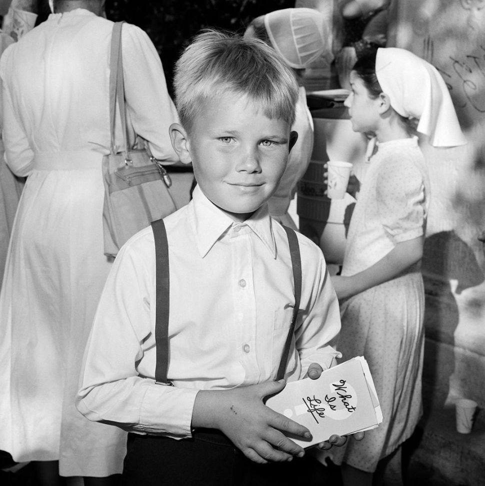 A black-and-white photograph shows a young boy in a buttoned shirt and suspenders holding a stack of cards while looking at the camera, with adults and children gathered in the background.