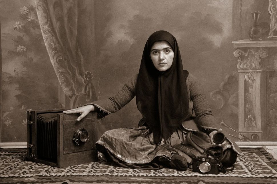 A young woman in traditional dress and headscarf sits on a patterned rug beside an antique large-format camera, holding a modern camera lens, posed against a painted studio backdrop evoking 19th-century portraiture.