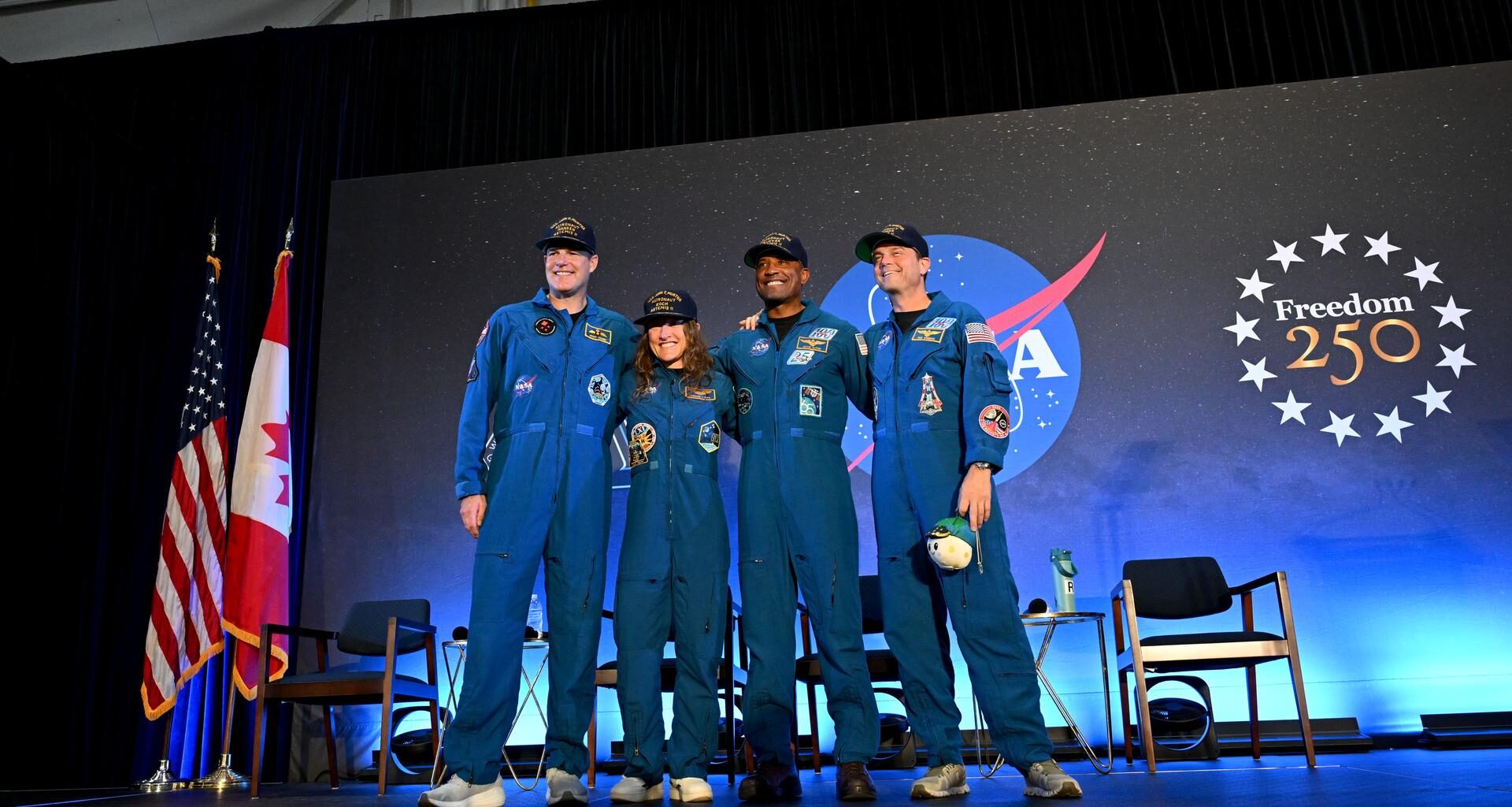 The four Artemis II crew members stand side-by-side and smiling in their blue flight suits on a stage at NASA Johnson. Bel