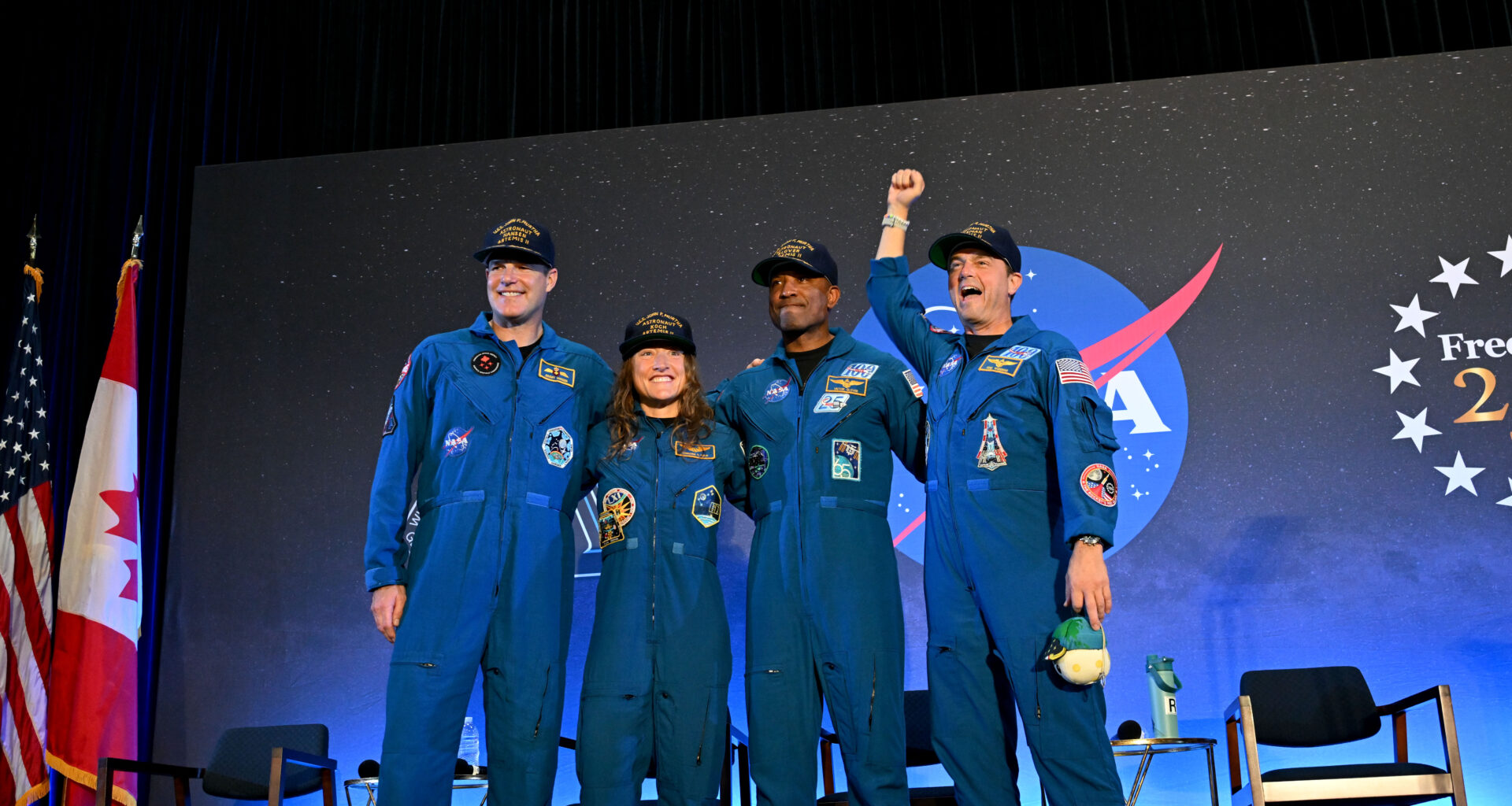 CSA (Canadian Space Agency) astronaut Jeremy Hansen and NASA astronauts Reid Wiseman, Victor Glover, and Christina Koch smile at the crowd during a news conference. They are all wearing blue jumpsuits with patches on the arms and chest areas. Wiseman raises his right fist in a victorious pose.