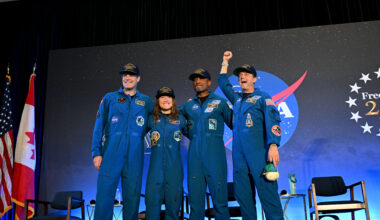 CSA (Canadian Space Agency) astronaut Jeremy Hansen and NASA astronauts Reid Wiseman, Victor Glover, and Christina Koch smile at the crowd during a news conference. They are all wearing blue jumpsuits with patches on the arms and chest areas. Wiseman raises his right fist in a victorious pose.