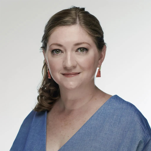 A woman with brown curly hair pulled back to one side looks at the camera, wearing red dangling earrings and a blue v-neck shirt