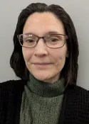A person with straight, dark brown hair and glasses is wearing a green turtleneck sweater and a black cardigan, posing in front of a plain light gray background.