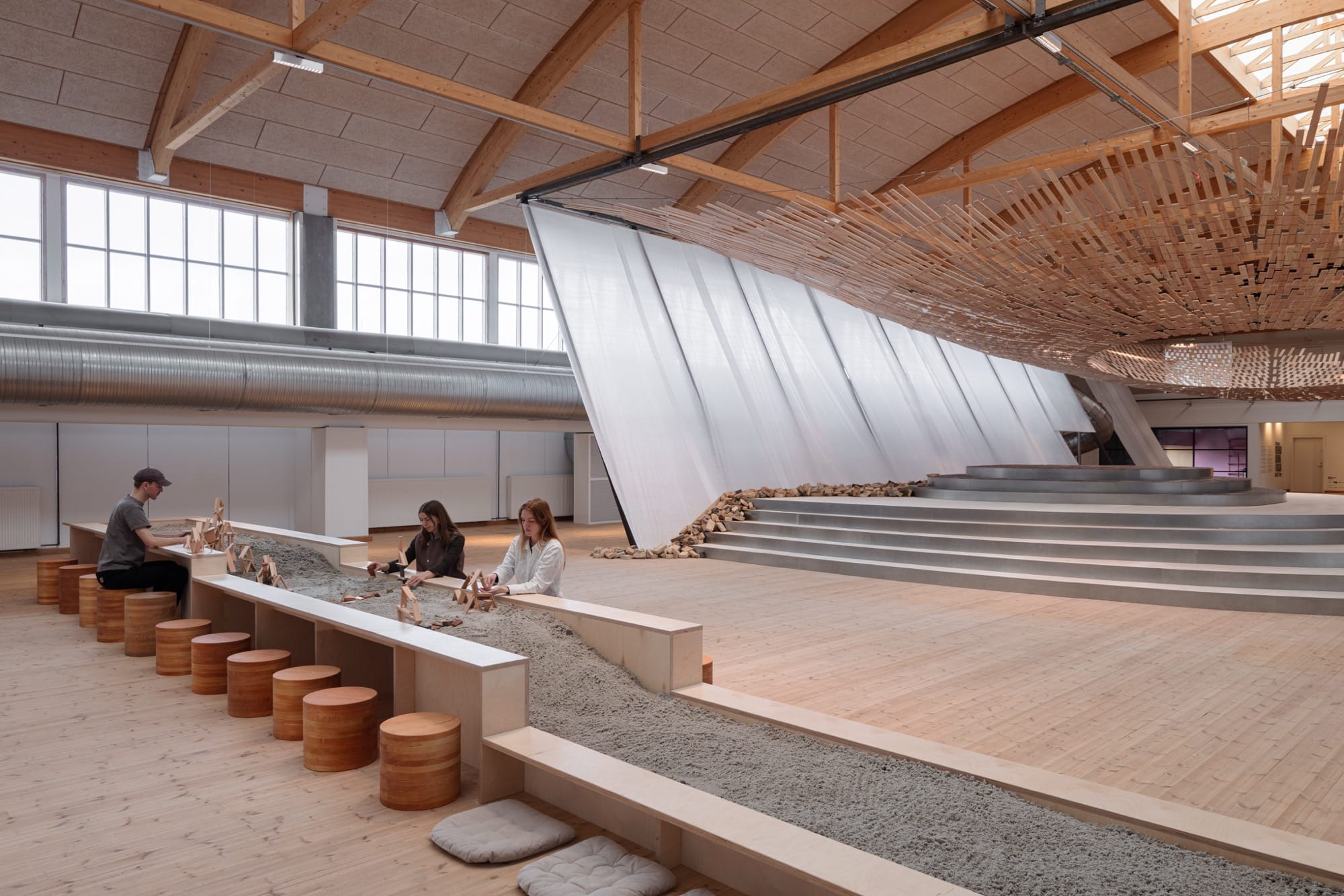 visitors work at a table and enjoy an installation in which light streams through a wooden canopy by Kengo Kuma